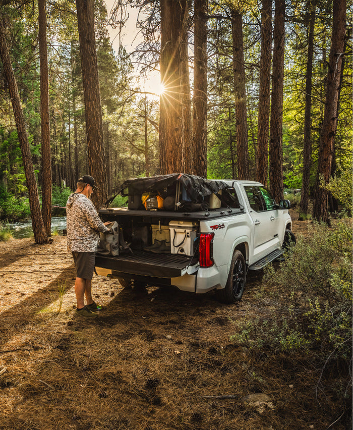 White Toyota Tundra pickup truck outfitted with a DiamondBack HD bed cover and Stronghold Cargo Net hauling overland gear in the woods.