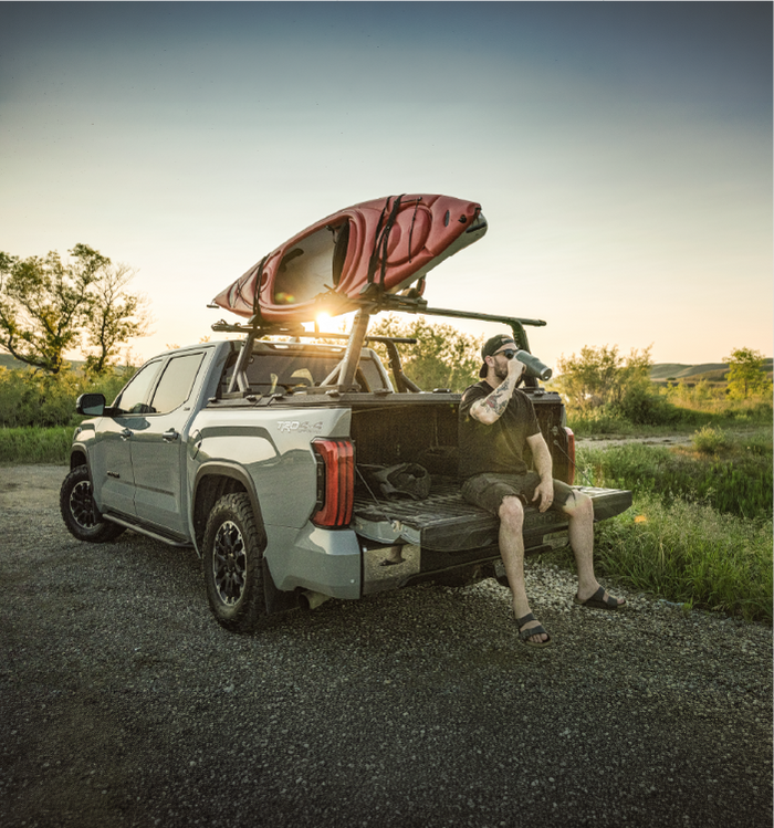 Man sitting on open tailgate of a Toyota pickup truck outfitted with a DiamondBack HD bed cover, Yakima OverHaul HD rack system, and a kayak loaded on top.